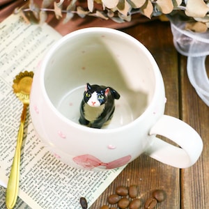 May include: White ceramic mug with a black and white cat figurine inside. The mug has a pink bow and pink polka dots on the outside.