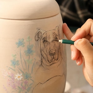 Puede incluir: Una urna de cer&aacute;mica de color beige claro con tapa, que presenta una ilustraci&oacute;n pintada a mano de la cara de un perro en tinta negra y un grupo de peque&ntilde;as y delicadas flores en tonos pastel azul, rosa y blanco en la superficie curva.