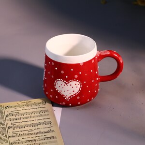May include: Red ceramic mug with white polka dots and a white heart design. The heart has white polka dots inside.