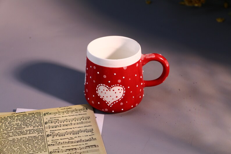 May include: Red ceramic mug with white polka dots and a white heart design. The heart has white polka dots inside.