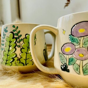May include: Two ceramic mugs with hand-painted designs. One mug features a green leafy pattern, while the other has a purple flower design with yellow centers.