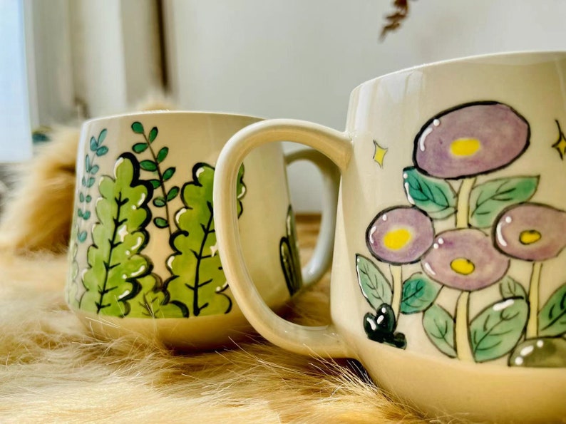 May include: Two ceramic mugs with hand-painted designs. One mug features a green leafy pattern, while the other has a purple flower design with yellow centers.