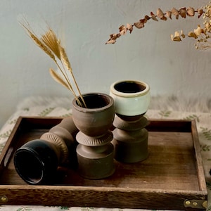 May include: Three brown ceramic candle holders with a white ceramic top on a wooden tray. The tray is on a white and green patterned surface. Dried wheat stalks are in one of the candle holders.