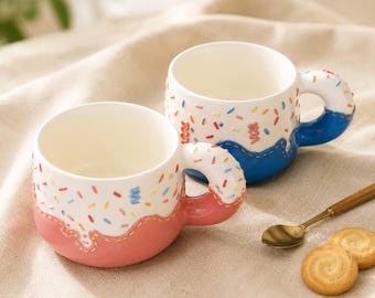 Peut inclure: Deux tasses en céramique avec une partie supérieure blanche parsemée de vermicelles colorés et une base unie rose ou bleue. Chaque tasse possède une anse en forme de beignet, la tasse rose ayant une base rose et la bleue une base bleue.