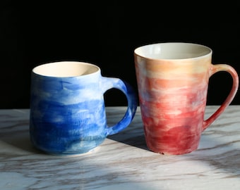 Op de afbeelding: Twee keramische mokken met een aquarel-achtig ontwerp. De ene mok is blauw en wit, de andere is rood en wit.