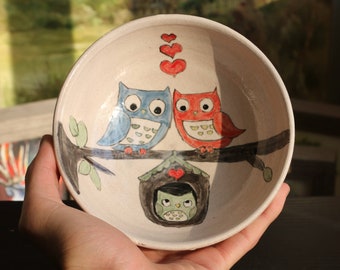 May include: A white ceramic bowl with a hand-painted design of two owls perched on a branch with hearts above them. The owls are blue and red, and the branch has green leaves. There is a small owl in a nest below the branch.