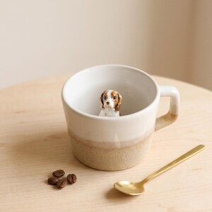 May include: A white ceramic mug with a brown and beige base and handle, containing a small dog figurine. A gold spoon and coffee beans sit beside the mug on a wooden surface. The dog figurine has brown and white markings.