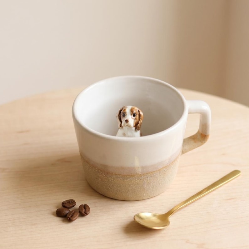 May include: A white ceramic mug with a brown and beige base and handle, containing a small dog figurine. A gold spoon and coffee beans sit beside the mug on a wooden surface. The dog figurine has brown and white markings.