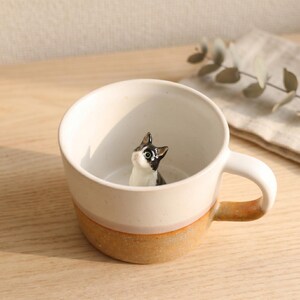 May include: A cream-coloured ceramic mug with a speckled tan base. A small, black and white cat figurine sits inside the mug. The handle is white with a tan accent. The mug is on a wooden surface.
