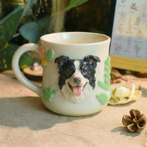 May include: Cream-colored ceramic mug featuring a 3D sculpted Border Collie dog face with black and white fur and a pink tongue. The mug is decorated with green leaves and flowers, and has a curved handle.