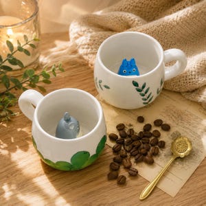 Op de afbeelding: Twee witte keramische mokken met handvatten, elk met een stripfiguur erin. Eén mok heeft een blauw karakter, de andere een grijs karakter. Groene blad- en wijnstokontwerpen versieren de mokken. Koffiebonen en een gouden lepel zijn ook aanwezig.