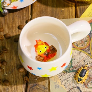 May include: A white ceramic mug with a handle, decorated with colorful stars. Inside the mug is a small, detailed figurine of a cartoon fire element. The mug is on a wooden surface with coffee beans scattered around.