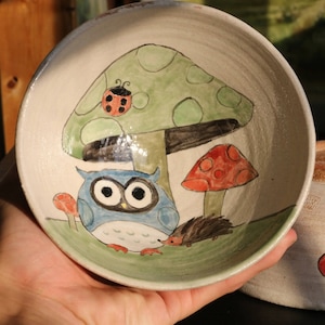 May include: Two ceramic bowls, one with a blue owl, a red mushroom, and a green mushroom with a ladybug on top, and the other with three red hearts.