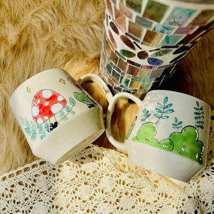 May include: Two ceramic mugs with hand-painted designs. One mug features a red and white mushroom with green leaves, the other has green leaves and a green hill.