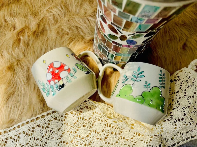 May include: Two ceramic mugs with hand-painted designs. One mug features a red and white mushroom with green leaves, the other has green leaves and a green hill.