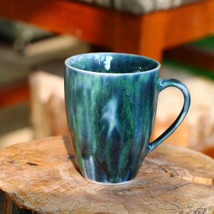 Puede incluir: Una taza de cerámica azul y verde con un esmalte marmoleado único. La taza tiene un cuerpo redondeado y un asa.