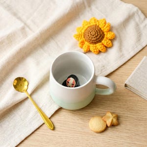 May include: A white ceramic mug with a green and white gradient, featuring a character figurine inside. A gold spoon, a crocheted sunflower, and cookies are arranged on a wooden surface with a linen cloth.