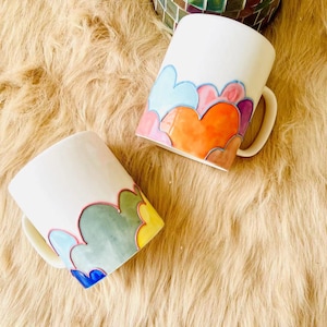 Pode incluir: Duas canecas de cerâmica branca com desenhos abstratos coloridos. As canecas têm uma alça do lado direito e são decoradas com uma fileira de nuvens estilizadas em tons de azul, verde, amarelo, laranja e rosa.
