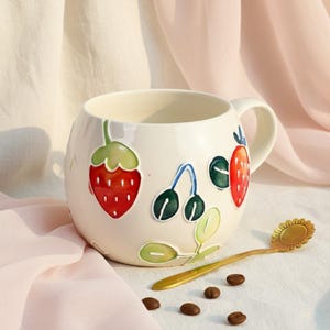 Puede incluir: Taza de cerámica color crema con asa, adornada con ilustraciones coloridas de fresas y cerezas. Una cuchara dorada con diseño de girasol descansa junto a la taza, con varios granos de café esparcidos cerca.