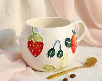 May include: Cream-colored ceramic mug with a handle, adorned with colorful illustrations of strawberries and cherries. A gold-colored spoon with a sunflower design rests beside the mug, with several coffee beans scattered nearby.