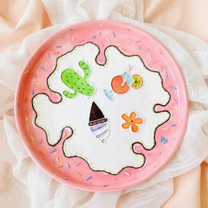 Può includere: Un piatto in ceramica rosa con un design a goccia bianco. Il piatto è decorato con illustrazioni colorate di un cactus, un cocktail, un cono gelato e un fiore. Il bordo è cosparso di puntini colorati.