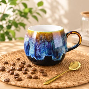 May include: A round ceramic mug featuring a reactive glaze with a gradient of amber, blue, and black colours. The mug has a curved handle and a white interior. A gold-toned spoon with a sunflower-shaped head rests beside the mug on a woven mat.