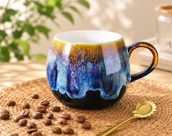 Peut inclure: Une tasse en céramique ronde avec un émail réactif présentant un dégradé de couleurs ambre, bleu et noir. La tasse possède une anse incurvée et un intérieur blanc. Une cuillère dorée en forme de tournesol repose à côté sur un tapis tressé.