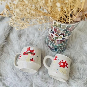May include: Two white ceramic mugs with mushroom designs. One mug features a red mushroom with white spots and the other has a red mushroom with a smiling face. Both mugs have a white background with colourful dots.