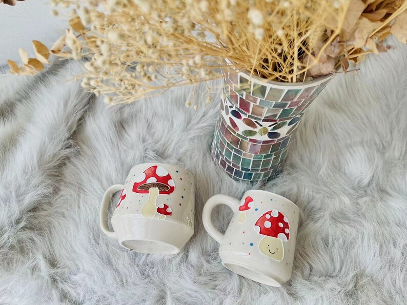 May include: Two white ceramic mugs with mushroom designs. One mug features a red mushroom with white spots and the other has a red mushroom with a smiling face. Both mugs have a white background with colourful dots.