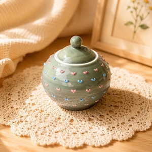 May include: A sage green ceramic jar with a lid, adorned with pink and blue heart-shaped embellishments. The jar sits on a cream-colored, crocheted doily. A small, round knob tops the lid.