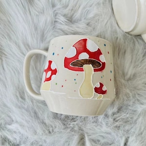 May include: A white ceramic mug with a red and white mushroom design. The mug has a handle and is decorated with colourful dots.