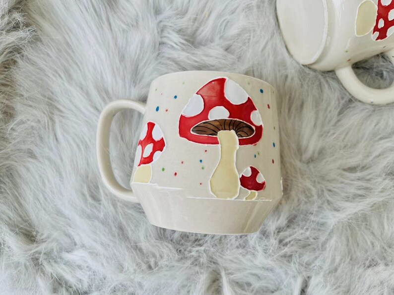 May include: A white ceramic mug with a red and white mushroom design. The mug has a handle and is decorated with colourful dots.