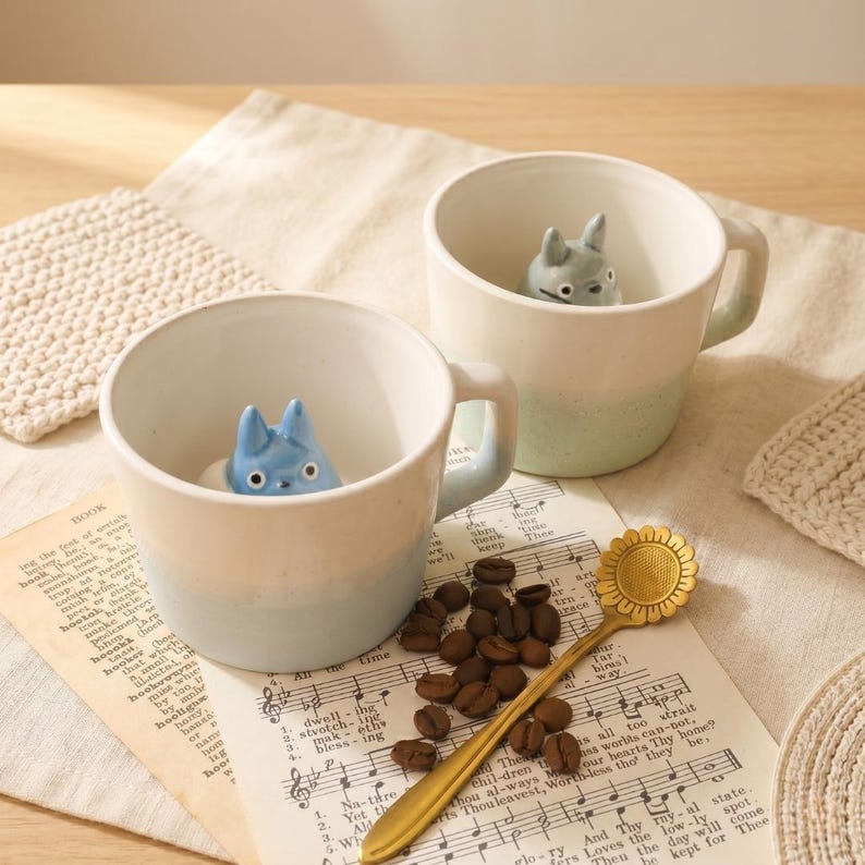 May include: Two ceramic mugs with a gradient colour scheme, each featuring a small character figurine inside. A gold-coloured spoon shaped like a sunflower and coffee beans are placed on sheet music. The mugs have light blue and green handles.