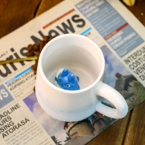May include: A white ceramic mug with a blue ombre design. The mug has a handle and a small blue figurine inside.