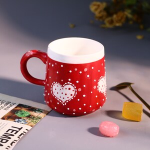 May include: Red ceramic mug with white polka dots and a white heart with polka dots. The mug has a white rim and a white handle.