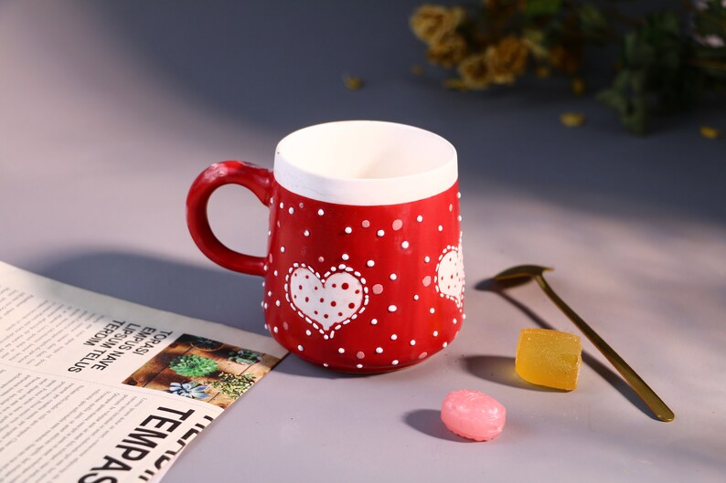 May include: Red ceramic mug with white polka dots and a white heart with polka dots. The mug has a white rim and a white handle.