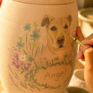 Puede incluir: Una urna de cer&aacute;mica de color tostado claro con un retrato pintado a mano de un perro marr&oacute;n rodeado de flores silvestres coloridas en tonos azules, morados y blancos. El nombre Angel est&aacute; pintado en letra cursiva debajo del perro con un pincel fino.
