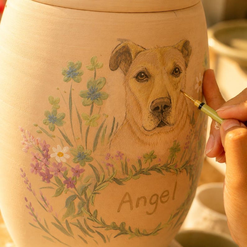 Puede incluir: Una urna de cer&aacute;mica de color tostado claro con un retrato pintado a mano de un perro marr&oacute;n rodeado de flores silvestres coloridas en tonos azules, morados y blancos. El nombre Angel est&aacute; pintado en letra cursiva debajo del perro con un pincel fino.