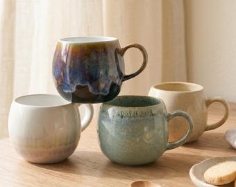 May include: Four ceramic mugs with rounded bodies and curved handles are arranged on a wooden surface. The mugs feature a variety of colors, including blue, green, and beige, with a glossy finish. A wooden spoon and a small plate with cookies are also visible.