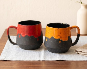Op de afbeelding: Twee keramische mokken met een uniek ontwerp. Eén mok is rood en de andere oranje, met een zwarte basis en handvat. De mokken hebben een gespikkelde textuur en een zwart interieur. Ze staan op een lichtgrijze doek op een houten tafel.