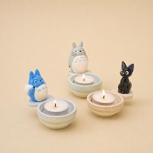 Peut inclure: Ensemble de bougeoirs en céramique représentant des personnages d'un film d'animation populaire. L'ensemble comprend une créature bleue, une figurine grise ressemblant à un lapin et un chat noir. Chaque personnage est posé sur un petit plat rond de couleur pastel contenant une bougie chauffe-plat allumée.