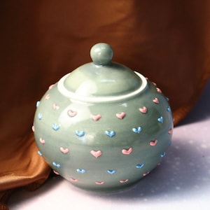 Puede incluir: Un azucarero de cerámica verde con tapa, decorado con corazones rosas y azules.