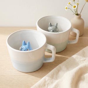 May include: Two ceramic mugs with a gradient colour scheme of white and blue-green. Each mug features a small, cartoonish animal figure inside. One is blue, and the other is grey. The mugs have a simple handle design and are set on a light-coloured surface.