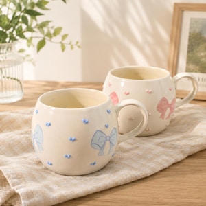 Puede incluir: Dos tazas de cerámica blanquecina con cuerpos y asas redondeadas. Una taza presenta diseños de lazo azul, la otra lazos rosas. Ambas tazas tienen pequeños detalles en forma de corazón. Las tazas están sobre un paño beige.