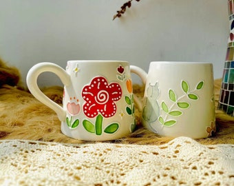May include: Two ceramic mugs with hand-painted designs. One mug features a red flower with green leaves and a white background. The other mug has a gray cat with green leaves on a white background.