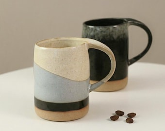 May include: Two ceramic mugs with large handles. One mug is cream, blue, black, and brown. The other mug is black and gray. Both mugs are on a white surface.