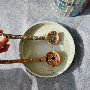 Puede incluir: Dos cucharas de madera con lunares coloridos pintados en ellas. Las cucharas están en un tazón de cerámica blanco.