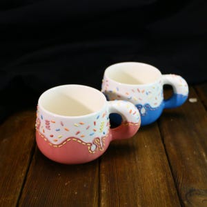 Op de afbeelding: Twee keramische mokken met een donutglazuurontwerp. De ene mok is roze met witte en blauwe sprinkles, de andere is blauw met witte en roze sprinkles.