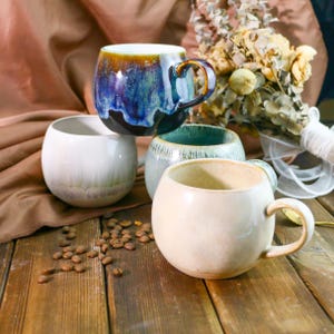 Könnte beinhalten: Vier Keramikbecher mit unterschiedlichen Glasuren. Die Becher sind rund und haben einen breiten Boden. Die Becher stehen auf einer Holzoberfläche mit Kaffeebohnen, die um sie herum verstreut sind.