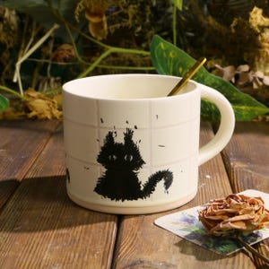 May include: Cream-colored ceramic mug with a black cat illustration. The mug has a grid pattern and a gold-colored spoon inside. The mug is on a wooden surface with a dried rose and a small card.
